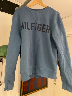 Blå sweatshirt från Tommy Hilfiger - Snygg blå sweatshirt från Tommy Hilfiger med tryckt logga över bröstet. Tröjan har rund halsringning, ribbade muddar och är tillverkad i mjukt bomullsmaterial. Perfekt för en avslappnad och stilren look.