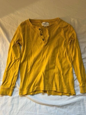 Gul långärmad henley från D.M.D för barn  - Gul långärmad henleytröja från D.M.D by denim day. Tröjan har rund halsringning med knappslå och tre knappar framtill. Tillverkad i mjukt bomullstyg, perfekt för en avslappnad och stilren look.