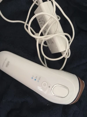 Braun Silk-expert 5 IPL hårborttagare - IPL hårborttagare från Braun i vit plast med bronsfärgad detalj. Ergonomisk form med tydliga knappar och indikatorlampor på ovansidan. Kommer med vit strömadapter och lång sladd. Perfekt för smidig hårborttagning hemma.