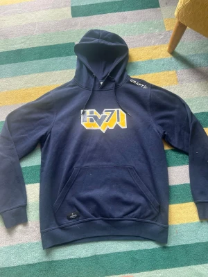 Blå HV71 hoodie från Craft - Mörkblå hoodie från Craft med HV71-logga i gult, vitt och blått på bröstet. Klassisk huva med snörning och stor magficka framtill. Perfekt för dig som vill representera HV71 med stil.