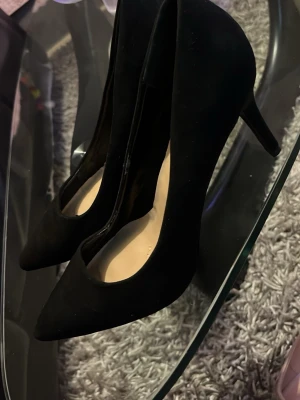 Svarta pumps från New Look - Säljer ett par klassiska svarta pumps från New Look med spetsig tå och smal klack. Skorna har en elegant siluett och är tillverkade i ett mockaliknande material. Perfekta för dig som vill ha en stilren och tidlös look.
