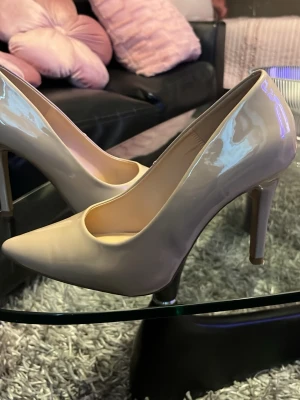 Beige lackpumps med spetsig tå - Snygga pumps i beige lack med spetsig tå och hög smal klack. Skorna har en klassisk och stilren design som passar till många olika outfits. Tillverkade i glansigt syntetmaterial för en trendig look.
