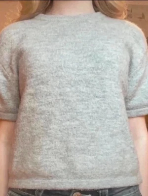 Ljusgrå stickad tröja Gina Tricot - Mysig ljusgrå stickad tröja från Gina Tricot med korta ärmar och rund hals. Tröjan har en enkel, rak passform och är tillverkad i ett mjukt, fluffigt material som känns skönt mot huden. Perfekt att styla med jeans eller kjol.