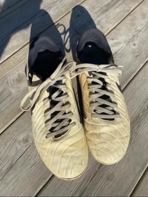 Beige Nike Tiempo fotbollsskor - Säljer ett par beige Nike Tiempo fotbollsskor med svart swoosh på sidan och svarta detaljer. Skorna har klassisk snörning och är tillverkade i syntetmaterial med mönstrad ovandel för bättre bollkontroll. Perfekta för dig som vill ha stilrena och funktionella skor på planen.