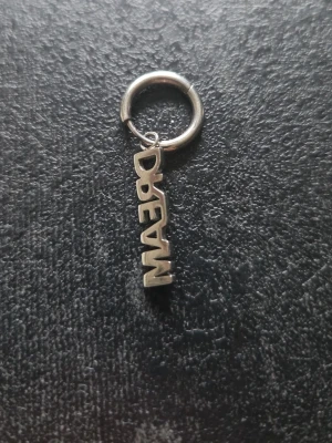 Silverörhänge med DREAM-hänge - Coolt örhänge i silverfärgad metall med ett hängande text-hänge där det står DREAM. Ringen är enkel och stilren, och texten ger en personlig touch. Perfekt för dig som vill ha något unikt och trendigt.