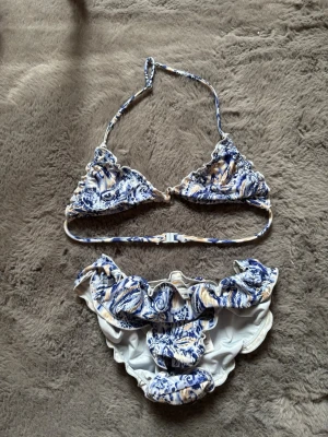 Blåvit volangbikini med mönster - Snygg bikini med blått och vitt mönster och volangdetaljer på både överdel och underdel. Triangelformad topp med knyt i nacken och klassisk bikiniunderdel. Perfekt för stranden eller poolen i sommar. De blir större än vad de ser ut när man har på sig dem.