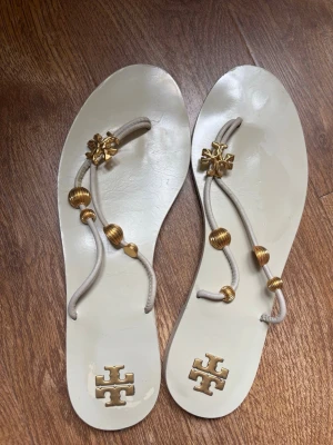 Beige sandaler från Tory Burch - Tory Burch platta flip-flop sandaler i elegant elfenbensvit/krämfärg med guldfärgade logotyper och pärldetaljer. Stilren minimalistisk design med bekväm platt sula och läderremmar. Begagnade med synliga bruksspår på sulorna och mindre skavanker, men fortfarande i fint skick med mycket användning kvar. Perfekta till sommaroutfits, strandsemester eller vardagsbruk.  Passform: Tory Burch-sandaler brukar vara små i storleken 
