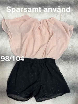 Ljusrosa och svart jumpsuit - Snygg jumpsuit med ljusrosa överdel och svarta shorts. Båda delarna har diskreta små prickar i tyget och resår i midjan för en bekväm passform. Perfekt för dig som gillar en luftig och trendig look.🌸 Sparsamt använd. Storlek 98/104