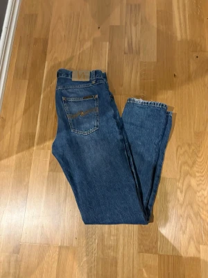 Blå jeans från Nudie Jeans - Klassiska blå jeans från Nudie Jeans med rak passform och femficksmodell. Jeansen har en mörkblå tvätt och Nudie-logga på bakfickan. Perfekta till vardags och gjorda i slitstarkt denimtyg.
