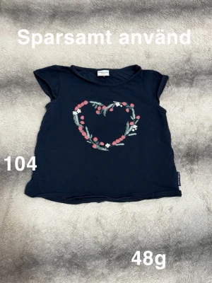 Mörkblå topp med hjärtmönster - Söt mörkblå topp från Polarn O. Pyret med korta ärmar och ett hjärta av blommor framtill i rosa, vitt och grönt. Perfekt för dig som gillar gulliga detaljer och mjuka material.🌸 Sparsamt använd. Storlek 104
