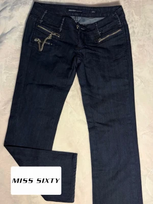 Mörkblå jeans Miss Sixty - Snygga mörkblå jeans från Miss Sixty med raka ben och coola dragkedjor samt kedjedetalj framtill. Klassisk femficksmodell med låg midja och diskreta sömmar. Perfekta för dig som gillar en edgy stil.