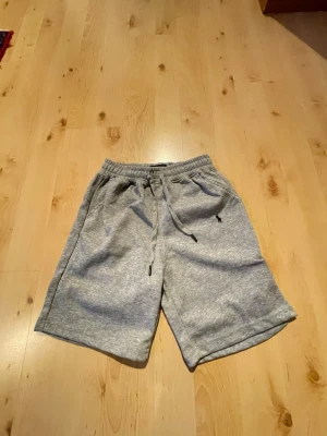 Grå sweatshorts från Polo Ralph Lauren - Snygga grå sweatshorts från Polo Ralph Lauren med dragsko i midjan och två sidofickor. Klassisk logga broderad på vänster sida. Perfekta för chill dagar eller när du vill ha en avslappnad stil.