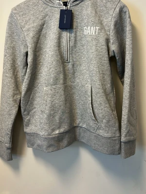 Grå hoodie från GANT med dragkedja - Snygg grå hoodie från GANT med halv dragkedja framtill och vit GANT-logga på bröstet. Huva med tryckt text längs kanten och stor magficka. Tillverkad i mjuk bomullsblandning, perfekt för en chill och avslappnad stil.