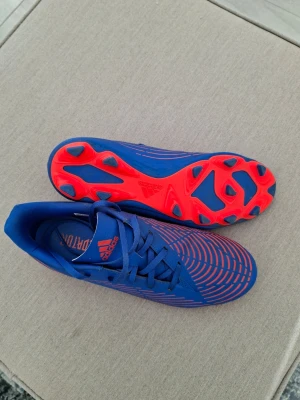 Adidas Predator blå fotbollsskor - Säljer ett par Adidas Predator fotbollsskor i blått med röda detaljer och mönster. Skorna har snörning, låg profil och dobbar i rött för bra grepp på planen. Snygg design med Predator-logga på sidan och ovandel i syntetmaterial.