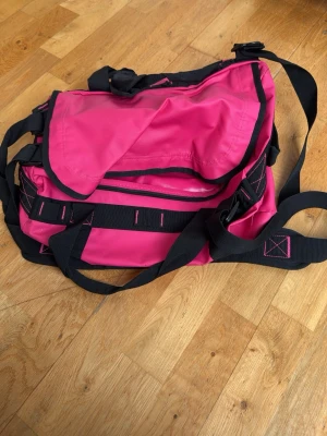 Rosa axelväska med svarta detaljer - Cool axelväska i starkt rosa nylon med svarta remmar och detaljer. Väskan har justerbar axelrem, flera fack och praktiska spännen. Perfekt för dig som vill sticka ut med en färgglad och funktionell väska.