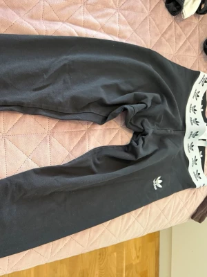 Svarta leggings från Adidas - Svarta leggings från Adidas med vit resår i midjan som har klassiska Adidas-loggor. Slimmad passform och stretchigt material som sitter skönt. Perfekta för en sportig look eller till chill-outfiten.