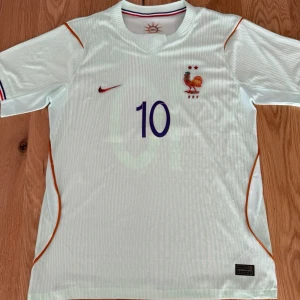 Frankrike fotbollströja Mbappé #10 - Vit fotbollströja från Nike med Frankrikes landslagsmärke, röda och blå detaljer samt nummer 10 och namnet Mbappé på ryggen. Tröjan har korta ärmar, rund hals och orangea linjer längs sidorna. Perfekt för fotbollsfans!