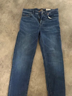 Blå slim fit jeans från boss  - Snygga blå slim fit jeans från boss med klassisk femficksdesign och dragkedjegylf. Jeansen har en lätt tvättad look och orangea detaljer vid midjan. Perfekta för en avslappnad stil.
