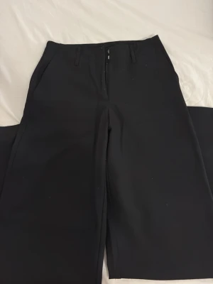 Svarta kostymbyxor från Vero Moda - Stilrena svarta kostymbyxor från Vero Moda med raka ben och hög midja. Byxorna har hällor för skärp och diskreta sidofickor. Perfekta för en clean och klassisk look. Materialet är mjukt och följsamt, vilket ger en snygg passform.
