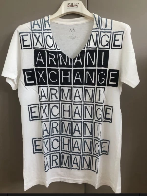 Vit t-shirt från Armani Exchange - Vit t-shirt från Armani Exchange med svart grafiskt tryck och stora bokstäver framtill. T-shirten har v-ringning och korta ärmar. Perfekt för en trendig och avslappnad look.