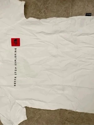 Vit t-shirt från The North Face - Vit t-shirt från The North Face med röd logga på bröstet och texten 'Never Stop Exploring' tryckt vertikalt. Klassisk passform och rund hals. Perfekt för en avslappnad och stilren look.