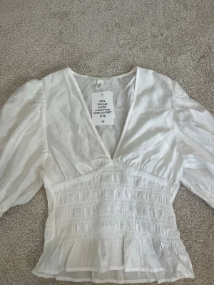 Vit blus med v-ringning från H&M - Superfin vit blus från H&M med djup v-ringning och smockad midja. Blusen har puffiga långärmar och är tillverkad i 89% viskos, delvis från Circulose. Perfekt till jeans eller kjol för en söt sommaroutfit.