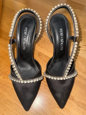 Svarta pumps med strass  - Snygga svarta pumps från Epe Shoes med spetsig tå och dekorerade med glittrande strass runt remmarna. Skorna har en klassisk klack och röd sula som ger en extra touch. Materialet är satinliknande och remmarna ger en elegant look.