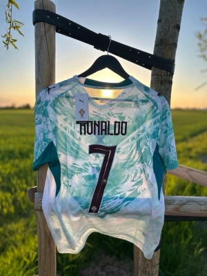 Portugal Ronaldo matchtröja Puma - Säljer en Portugal matchtröja från Puma med Ronaldo och nummer 7 på ryggen. Tröjan är vit med gröna och turkosa mönster, har korta ärmar och rund hals. Perfekt för fotbollsfans som vill sticka ut på planen eller läktaren.