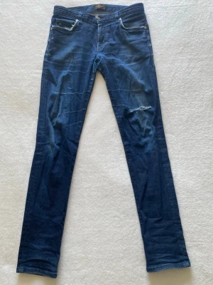 Mörkblå slim jeans - Säljer ett par mörkblå J Lindeberg jeans – modell Jay Mid-Rise / Slim Fit.Storlek W33/L34. Jeansen har en snygg slim fit-passform och är i väldigt bra skick och har en slitning vid knät för en vintage look. Bekväma och stilrena jeans som passar till det mesta.