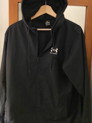 Svart vindjacka från Under Armour - Svart vindjacka från Under Armour med dragkedja framtill, huva med dragsko och vit logga på bröstet. Jackan är lätt och har meshfoder på insidan för extra komfort. Perfekt för blåsiga dagar och sportiga outfits.