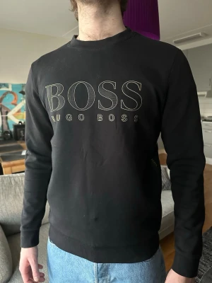 Hugo Boss sweatshirt - Säljer en sweatshirt från Hugo Boss med stor logga framtill i guld och dekorativa band med logga längs sidorna. Tröjan har rund halsringning, ribbade muddar och är tillverkad i mjukt material. Storlek M💫
