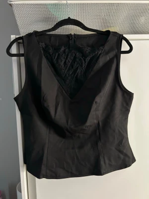 Svart ärmlös topp med spetsdetalj - Snygg svart ärmlös topp med v-ringning och dekorativ spets framtill. Toppens material känns slätt och har en figurnära passform som framhäver siluetten. Perfekt att styla med jeans eller kjol för en trendig look.
