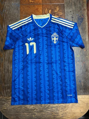 Sverige VM Borta fotbollströja Adidas - Säljer en blå fotbollströja från Adidas med Sveriges landslagsemblem och nummer 17 på bröstet. Tröjan har gula detaljer, klassiska Adidas-ränder på axlarna och namnet Gyökeres på ryggen. Tillverkad i lätt och ventilerande material, perfekt för träning eller match. Sprillans ny, har endast tagit bort prislappen.