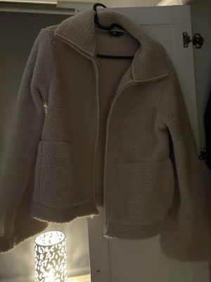 Beige teddyjacka med dragkedja - Mysig beige teddyjacka med hög krage och dragkedja framtill. Jackan har två stora fickor på sidorna och är tillverkad i fluffigt teddy-material som håller dig varm. Perfekt för kyliga dagar och enkel att matcha med olika outfits.