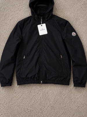 Svart vindjacka från Moncler - Snygg svart vindjacka från Moncler med huva och dragkedja framtill. Jackan har två fickor med dragkedja och Moncler-logga på ena ärmen. Tillverkad i lätt och slitstarkt material, perfekt för blåsiga dagar.