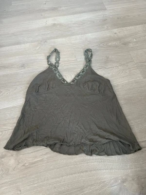 Khaki  linnetopp med spets från Lea L - Supersnygg khaki linnetopp från Lea L med dekorativa spetsband och pärldetaljer. Toppen har v-ringning och är ärmlös, perfekt för en trendig och avslappnad look. Lätt och luftig känsla, passar perfekt till jeans eller kjol.