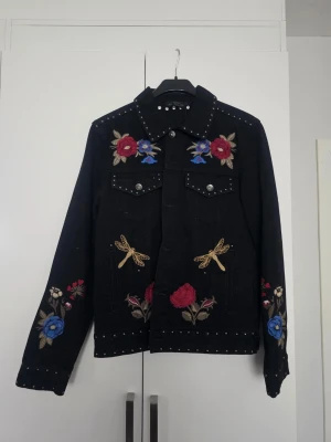 Svart jeansjacka med broderi och nitar - Nypris ca 800 kr. Ej använd. Snygg svart jeansjacka med färgglada broderade blommor och trollsländor på bröstet, ärmarna och nedtill. Jackan har silverfärgade nitar längs krage, fickor och ärmslut för en extra edgy look. Perfekt för dig som vill sticka ut med detaljer.