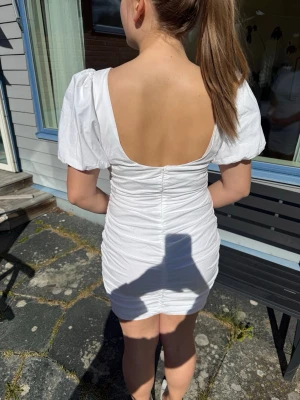 Vit kort puffärmad klänning - Snygg vit kort klänning med puffärmar och öppen rygg. Klänningen har en figurnära passform med snygg rynkad detalj längst hela ryggen. Den har en jättefin skärning runt halsen och sitter fint över hela kroppen. Perfekt till studenten, skolavslutningen eller midsommar. Använd 1 gång och är helt utan fläckar eller tecken på användning. 💞