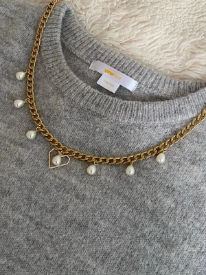 Guldfärgat halsband med pärlor - Trendigt halsband i guldfärgad rostfritt med hängande vita pärlor och en tunn hjärtformad detalj. Kedjan är chunky och ger en cool kontrast till de klassiska pärlorna. Perfekt accessoar för att lyfta din outfit.