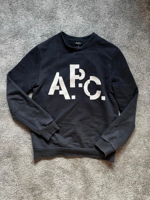 Svart sweatshirt från A.P.C. - Cool svart sweatshirt från A.P.C. med stor vit logga framtill. Tröjan har rund halsringning, långa ärmar och ribbade muddar vid ärmslut och nederkant. Perfekt för en avslappnad och stilren look.