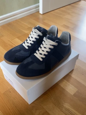 Maison Margiela navy sneakers - Snygga navyblå sneakers från Maison Margiela med klassisk siluett, vita snören och gummisula. Ovandelen är i mocka och läder för en lyxig känsla. Perfekta för dig som gillar stilrena och tidlösa sneakers med premiumkänsla.