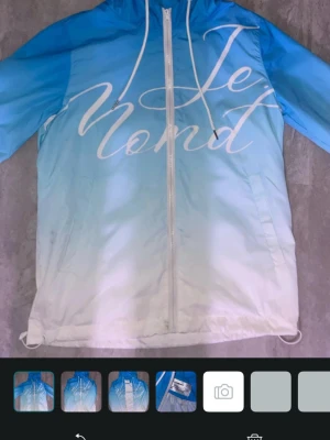 Blåvit vindjacka Jemond - Snygg vindjacka i blått och vitt med gradient effekt och stor vit text på framsidan. Jackan har huva med dragsko, hel dragkedja och två sidofickor. Perfekt för blåsiga dagar och streetwear-stil. Pris kan diskuteras