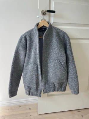 Grå teddyjacka från Vero Moda - Mysig grå teddyjacka från Vero Moda med två stora framfickor och ståkrage. Jackan har en rak passform och elastisk nederkant. Perfekt för kyliga dagar när du vill ha både stil och komfort.