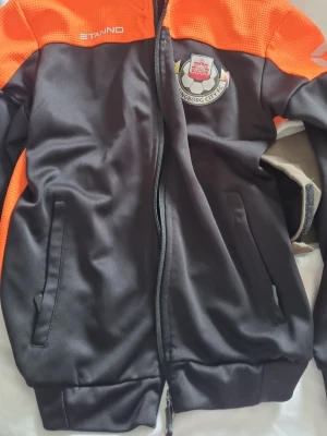 Svart och orange träningsjacka Stanno - Sportig träningsjacka i svart med orange axelpartier från Stanno. Jackan har dragkedja framtill, två fickor och klubbmärke för Helsingborg City FC på bröstet. Tillverkad i ett slätt, syntetiskt material som passar perfekt till träning.