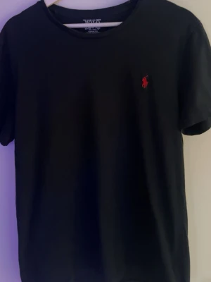 T shirt Ralph lauren - Svart t-shirt från Ralph Lauren med klassisk rund hals och korta ärmar. T-shirten har en röd broderad logga på bröstet och är tillverkad i mjuk bomull för en skön känsla.