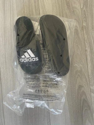 Svarta Adidas badtofflor - Svarta sandaler från Adidas med klassisk vit logga på ovandelen. Tillverkade i syntetmaterial och perfekta för att glida runt i hemma eller på stranden. Snygg och enkel design som passar till det mesta. PRISET KAN DISKUTERAS 