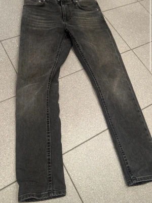 Svarta jeans från Nudie Jeans - Snygga svarta jeans från Nudie Jeans med klassisk femficksdesign och diskret slitning. Jeansen har slim ben och ikonisk brodyr på bakfickan. Perfekta för en stilren och avslappnad look.