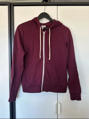 Vinröd hoodie med dragkedja - Vinröd hoodie med vit dragkedja och matchande vita snören i huvan. Tröjan har två fickor framtill och ribbade muddar vid ärmslut och nederkant. Perfekt för en avslappnad och stilren look.