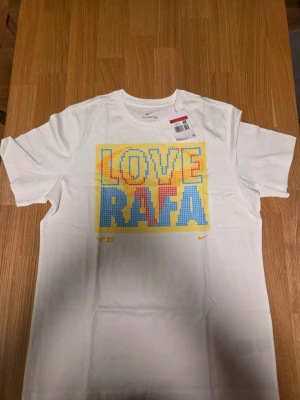 Nike vit t-shirt, Tennis , storlek L - Produktnummer fn5318-100  Vit t-shirt med trycket "LOVE RAFA" i pixelerad stil. T-shirten har en klassisk passform och korta ärmar. Det finns en liten Nike-logga broderad på bröstet och en "23" tryckt nedanför huvudtrycket.