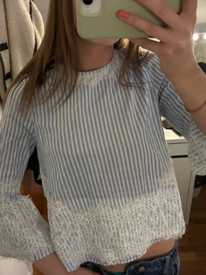 Blåvit randig blus med spetsdetaljer - Söt blus med blå och vita ränder, långärmad och dekorerad med vit spets nertill och vid ärmslut. Blusen har en öppen rygg med knytband i nacken för en extra detalj. Perfekt för dig som gillar romantisk och trendig stil.
